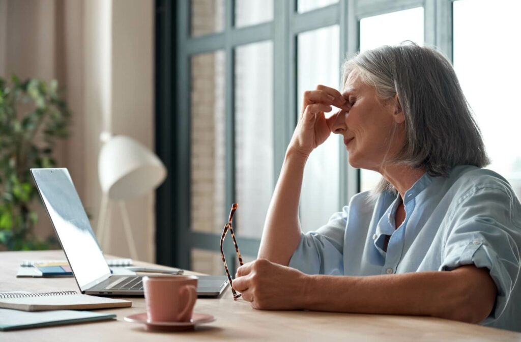 A person sits at a desk in front of a computer, holding a pair of reading glasses in their left hand while rubbing their eyes with their right due to eye strain.