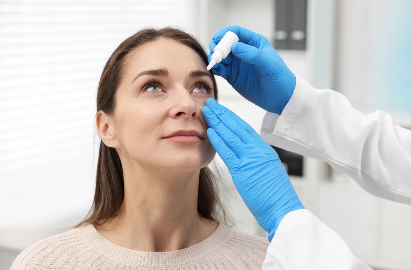 An optometrist wearing blue disposable gloves places eye drops into a patient’s eye.