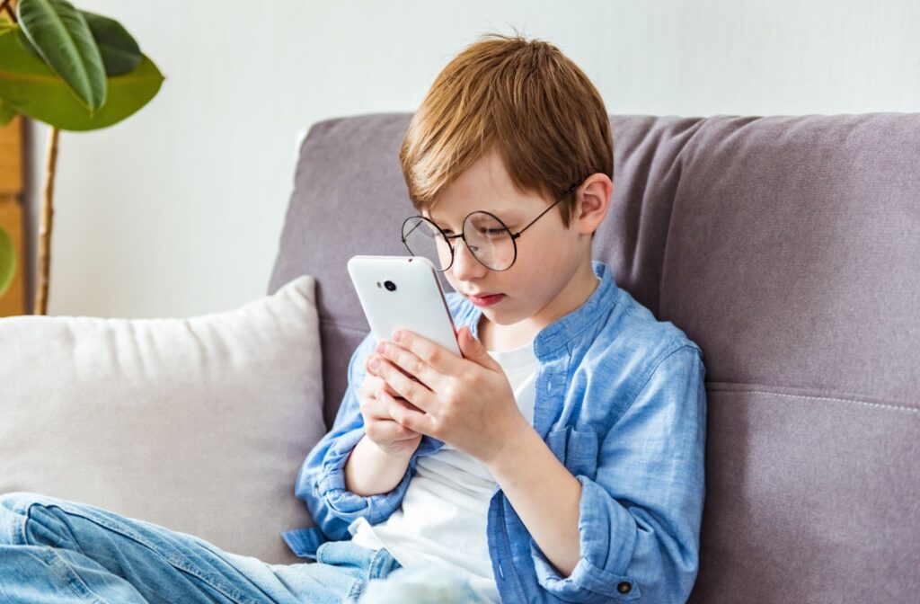 A young boy with myopia wearing glasses sits on the couch, holding his phone too close to his face.